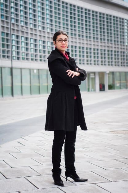 A woman standing confidently and facing forward. She is wearing a fitted blazer, dark jeans and ankle boots.